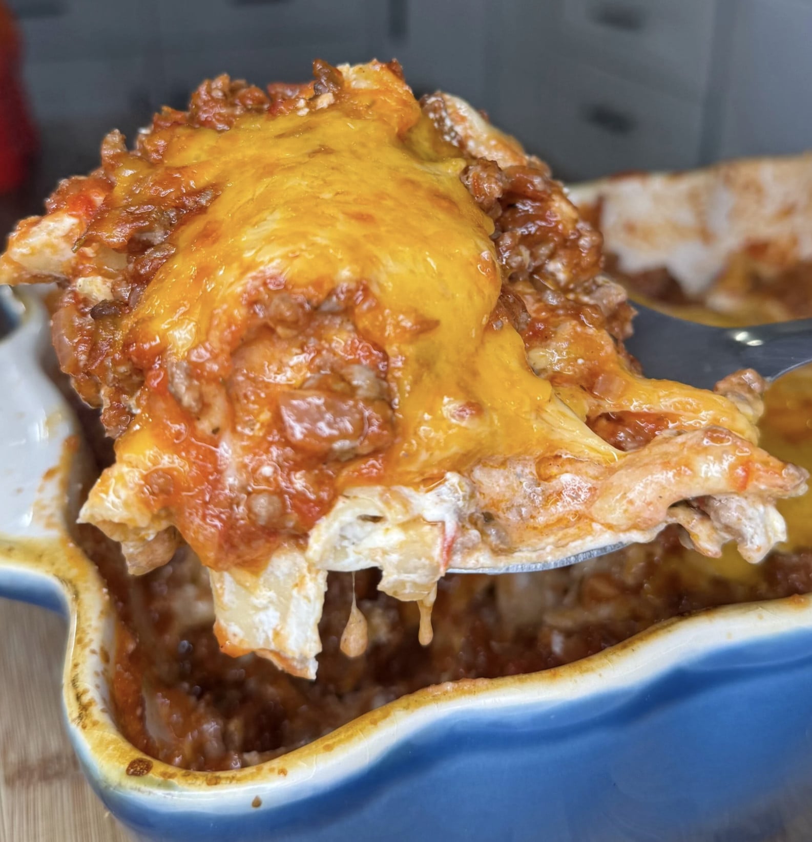 A metal spatula lifts a scoop of cheesy lasagna with meat sauce from a blue baking dish.