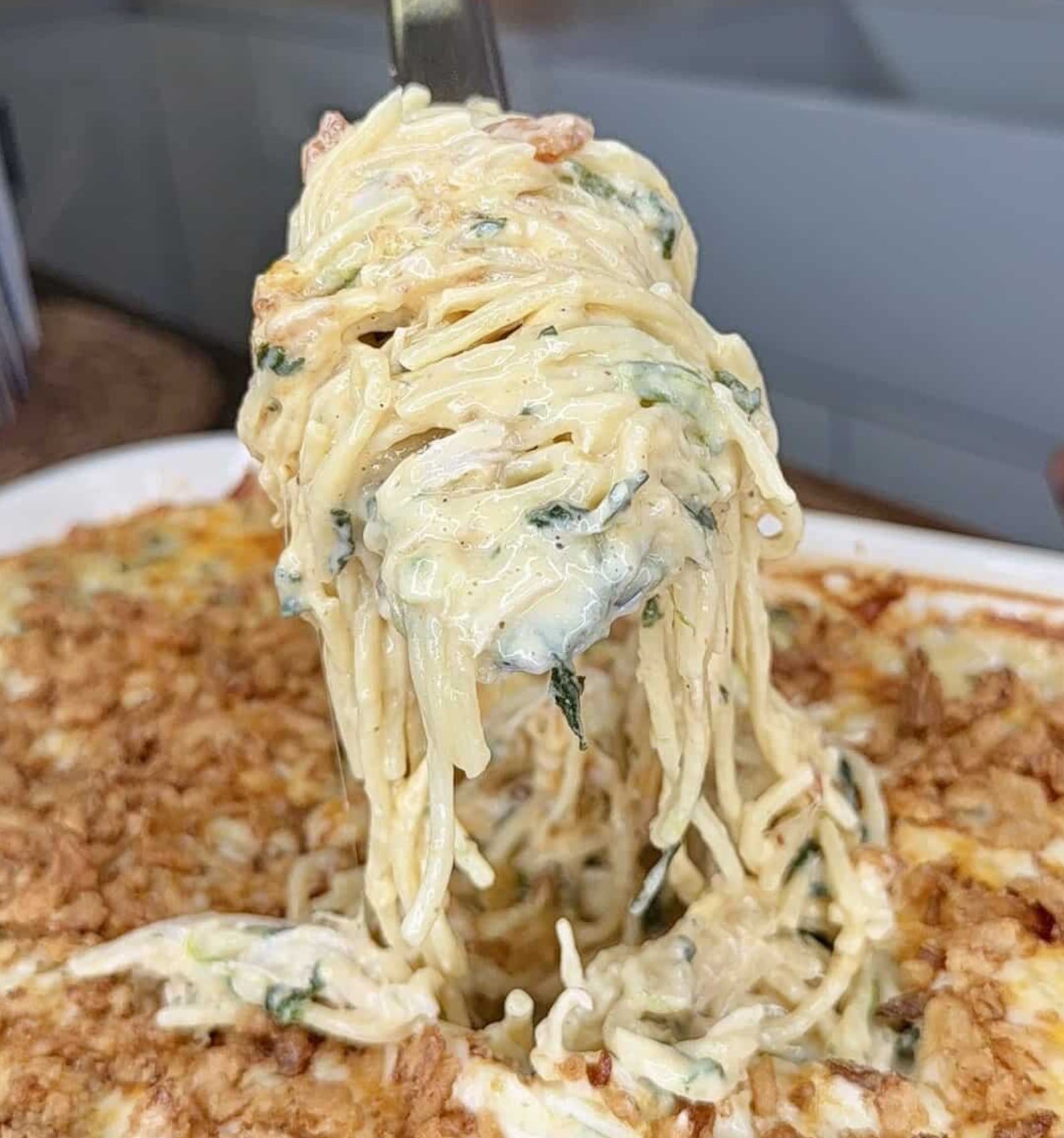 A fork lifts a portion of creamy pasta with spinach from a baked casserole topped with breadcrumbs.