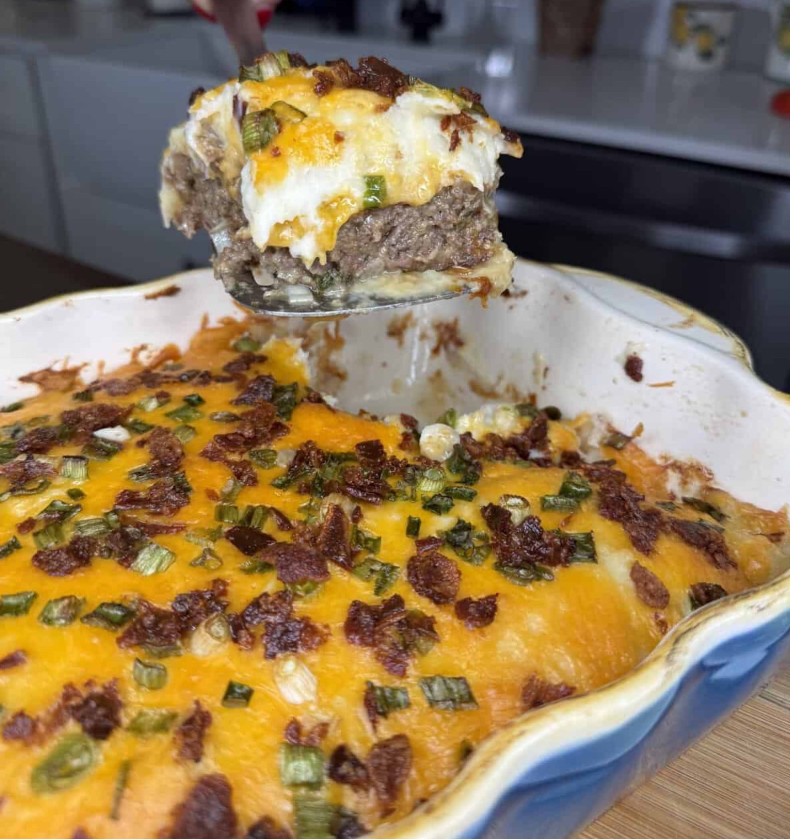 A metal spatula lifts a scoop of loaded baked potato casserole topped with melted cheese, bacon bits, and green onions from a baking dish.