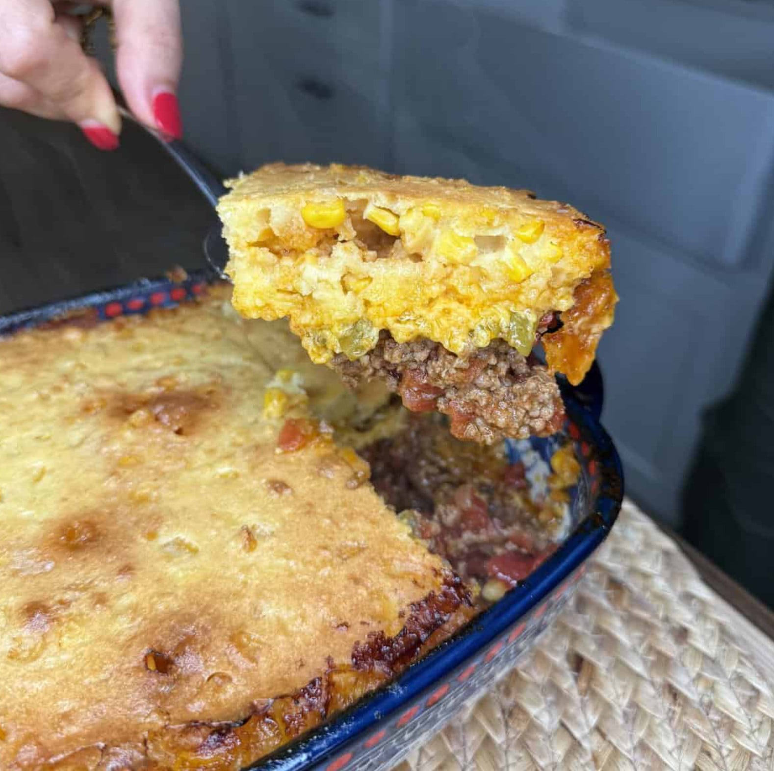 A serving spoon lifts a square of cornbread casserole, showing a layer of seasoned ground meat topped with golden cornbread mixed with corn kernels.