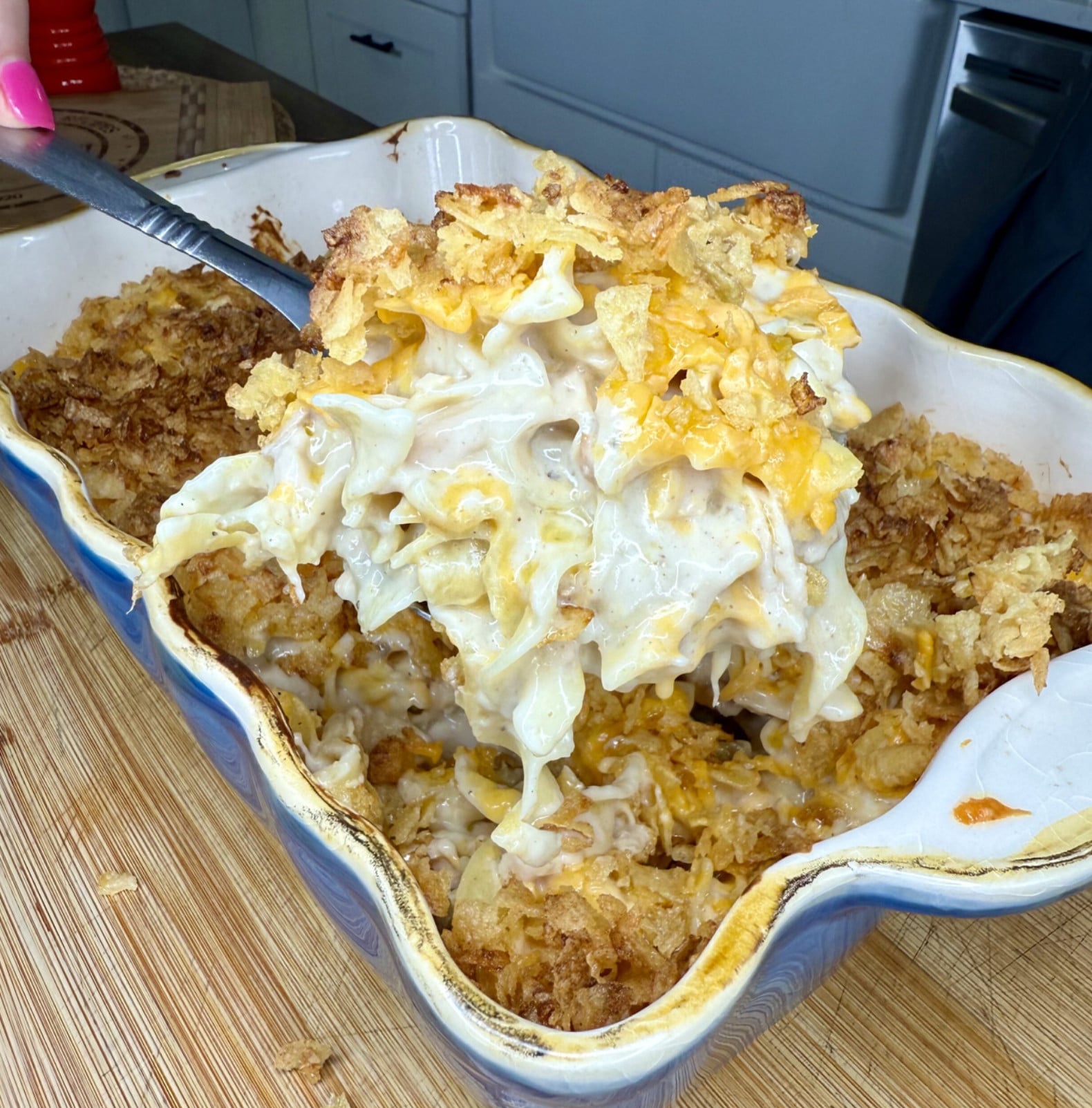 A metal serving spoon lifting a portion of creamy cheesy noodle casserole topped with crushed chips from a ceramic baking dish.