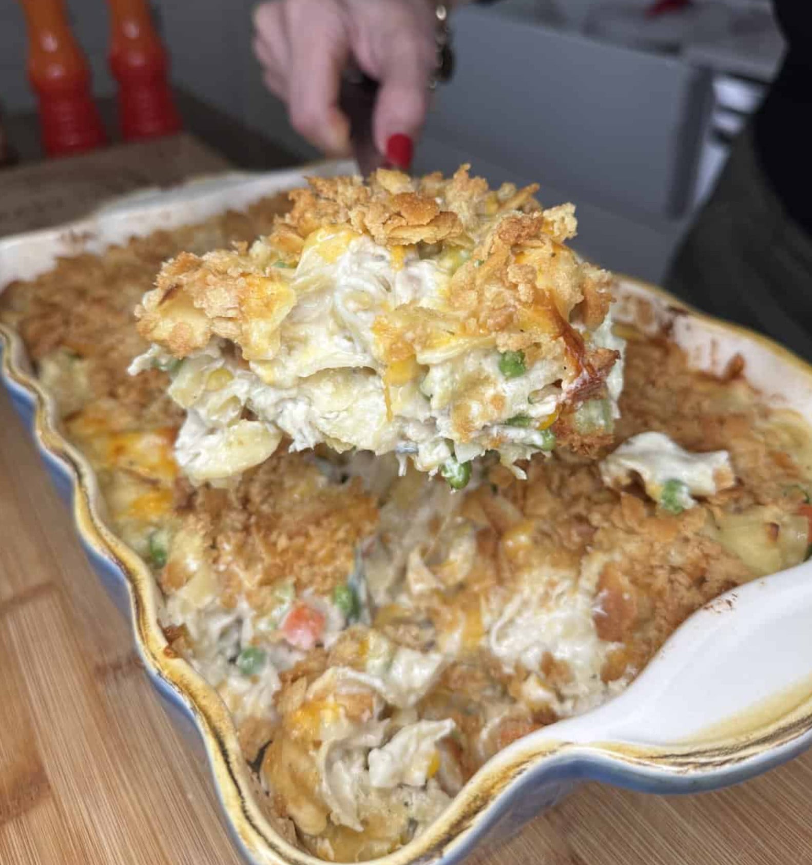 A hand uses a spatula to lift a large scoop of cheesy chicken casserole with a crispy topping out of a baking dish.