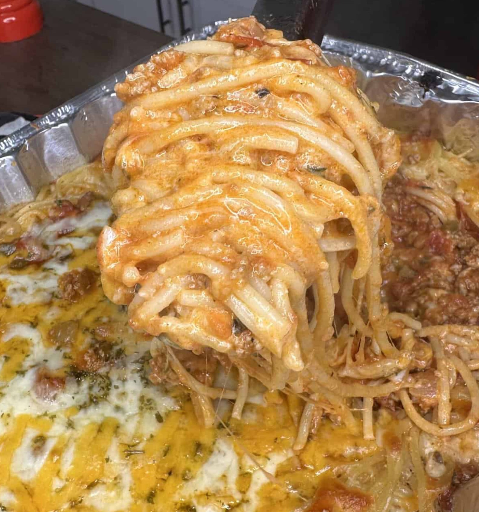 A serving spoon lifting a portion of spaghetti in a creamy meat sauce from a foil pan of baked pasta topped with melted cheese.
