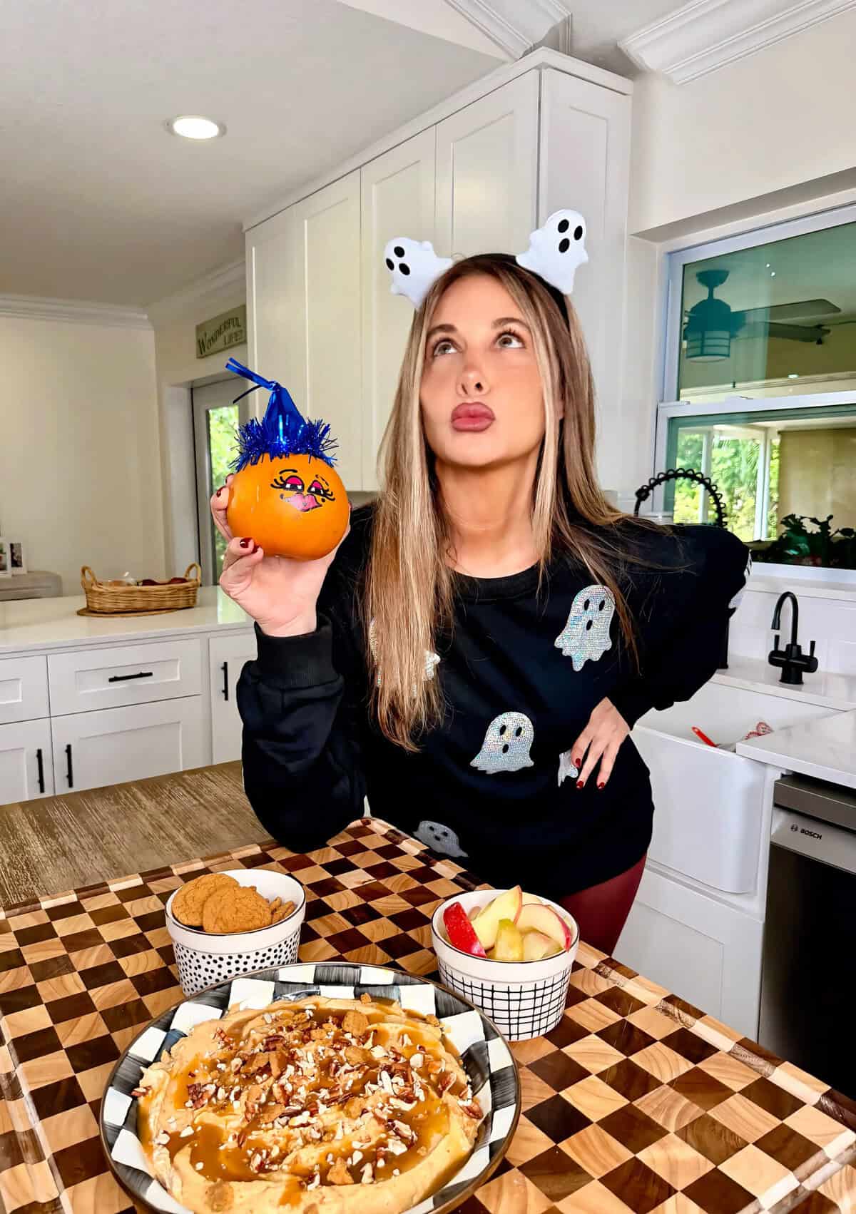 A woman wearing a ghost headband and a ghost-patterned sweater holds a small pumpkin decorated with a blue party hat, standing behind a table with a caramel-topped dessert, apple slices, and cookies.