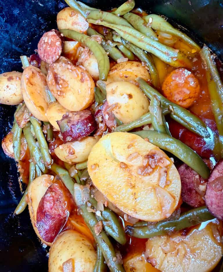 A slow cooker filled with sliced sausage, halved potatoes, and green beans in a light sauce.