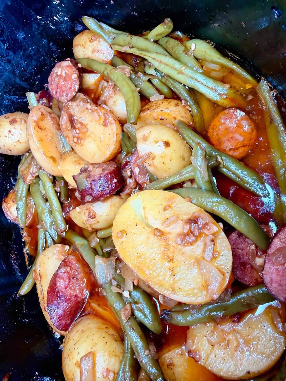 A slow cooker filled with a mixture of sliced sausage, potato wedges, and green beans in a savory sauce.