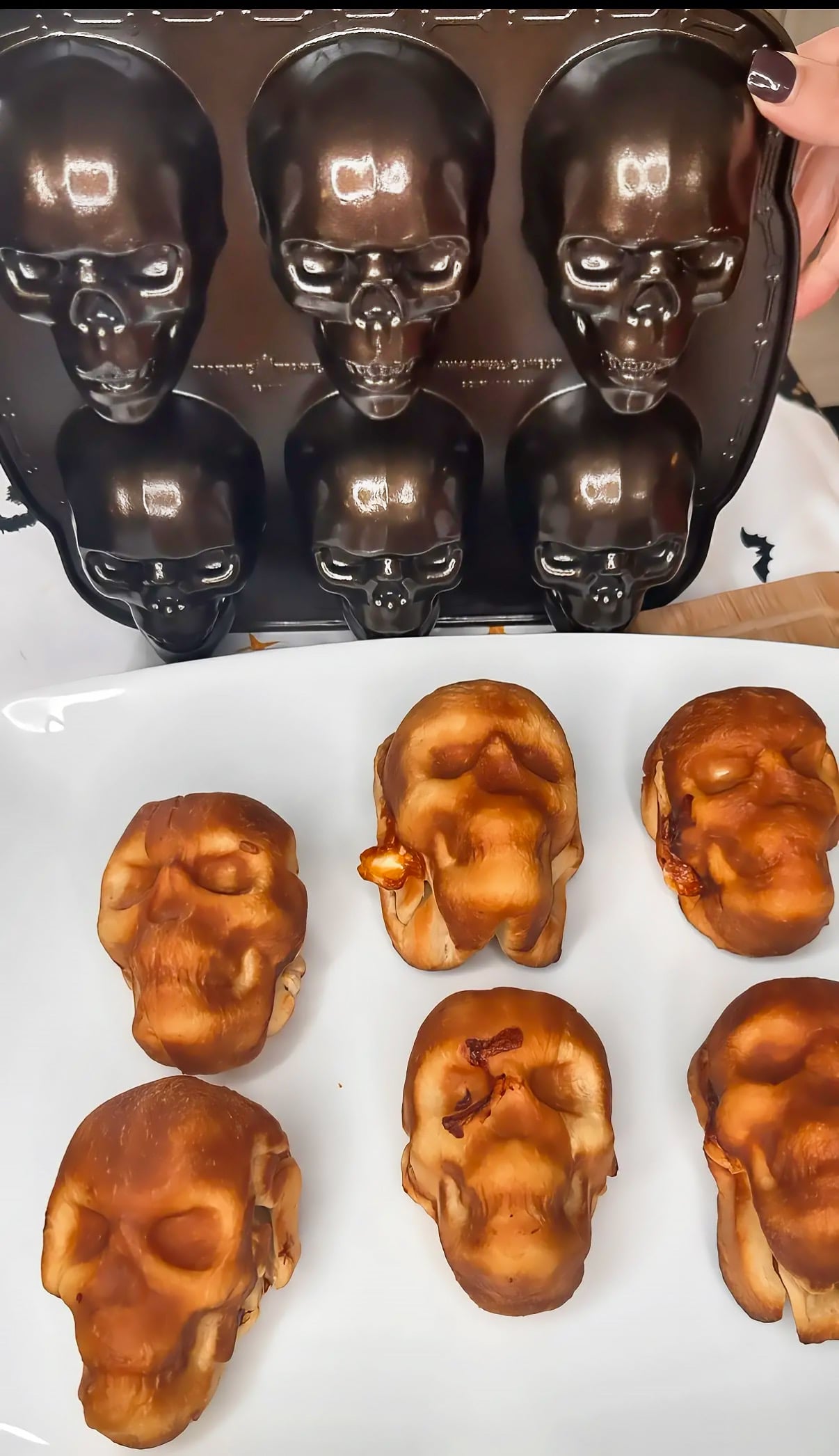 A hand holds a metal skull-shaped baking pan above a white platter containing six baked skull-shaped pastries.