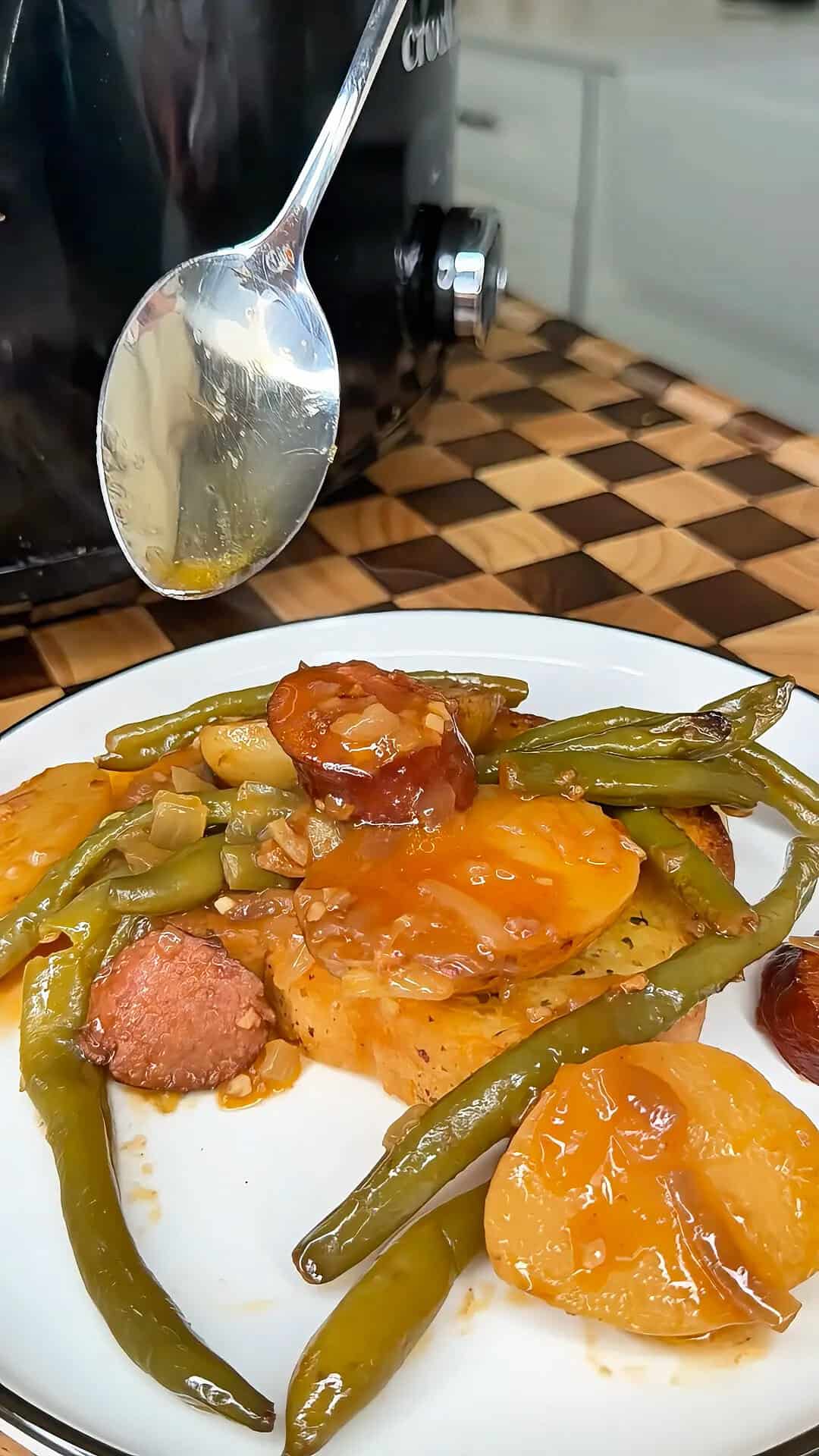 A spoon pours sauce over a plate of sliced sausage, green beans, and potatoes.