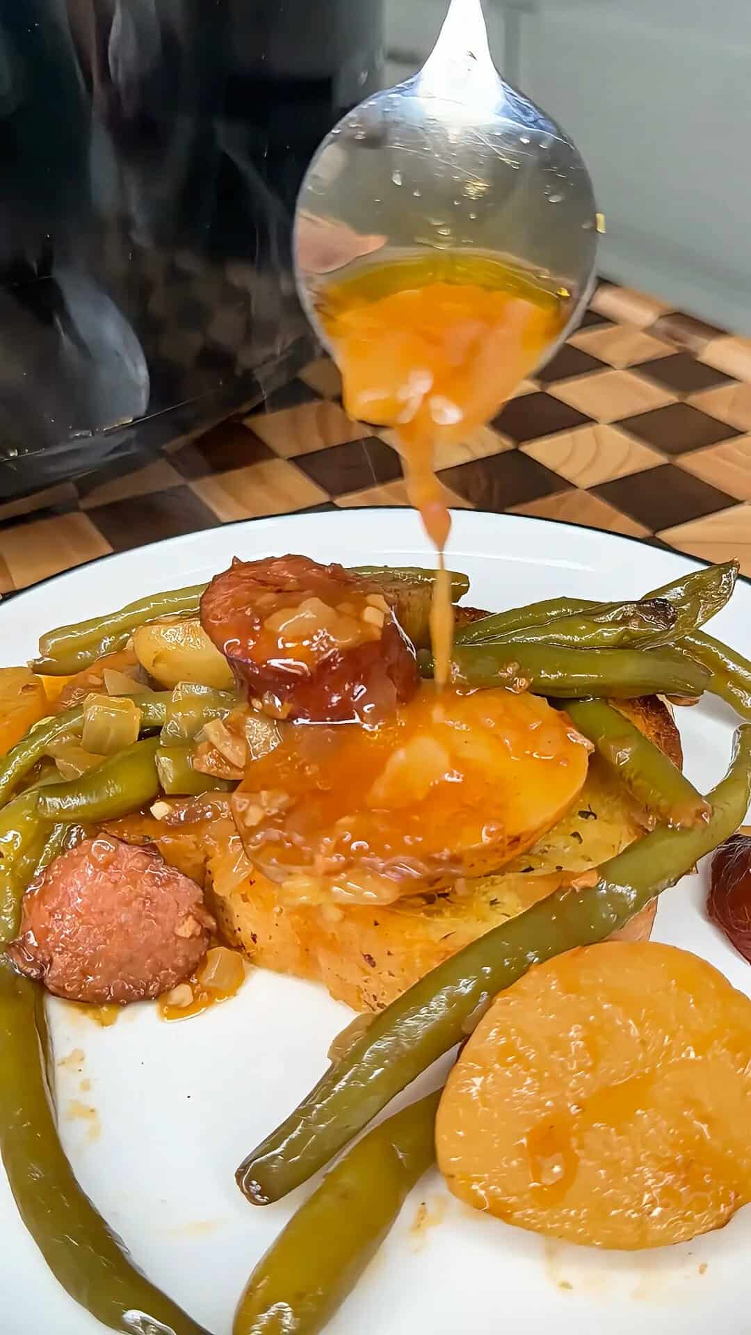 A spoon pours sauce over a plate of sliced sausage, green beans, and potatoes.